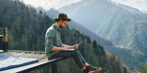 Young man freelancer traveler wearing hat anywhere working online using laptop and enjoying mountains view
